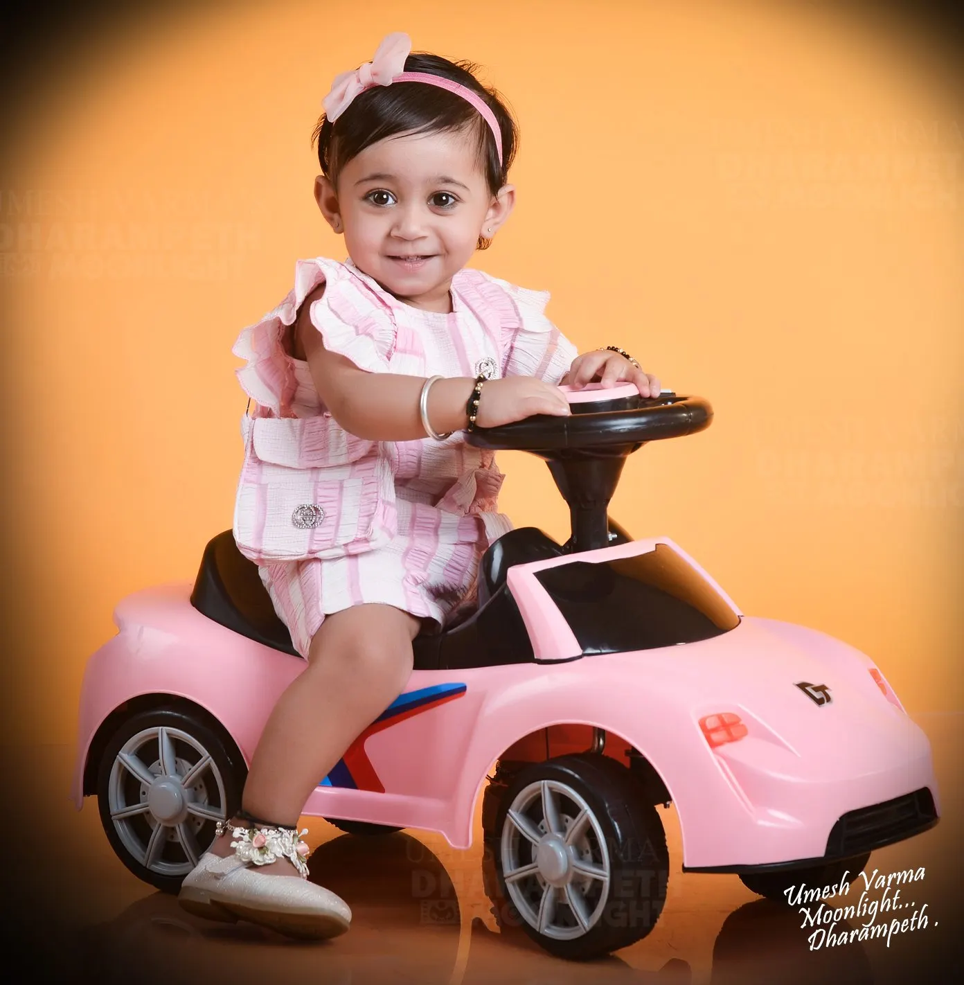 Toddler in a pink dress riding a toy car against an orange backdrop